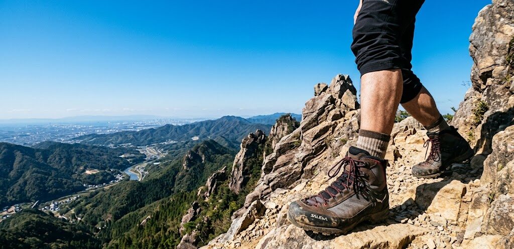 澄み切った青空の下、険しい岩山の登山道にしっかりと踏み出された丈夫なトレッキングシューズの足元。広大な風景を見下ろす、力強さと前進を感じさせるダイナミックな広角ショット。