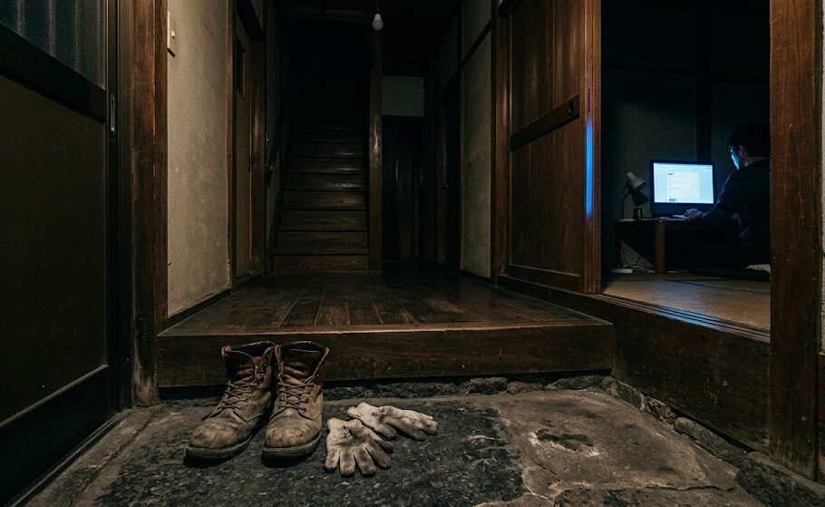 使い古された安全靴と作業用手袋が置かれた玄関の土間。その奥の部屋のわずかに開いたドアの隙間から、夜遅くにブログを執筆しているであろうパソコンのモニターの青い光がうっすらと漏れている、情景豊かな引きの実写風画像。