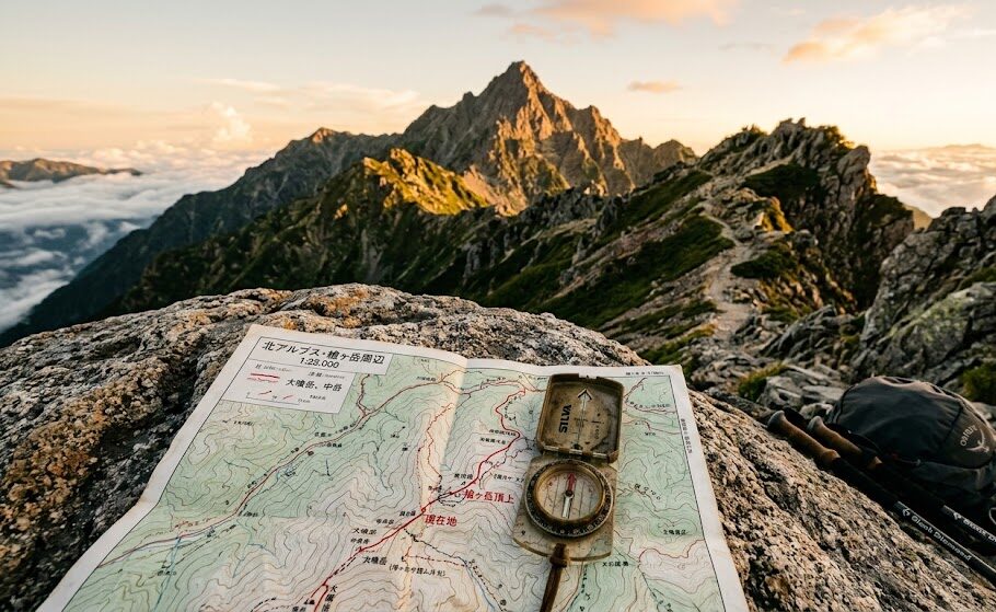 朝日が照らす険しくも美しい山道。その手前の岩の上に、使い込まれたコンパスと広げられた白地図が置かれている。誰の案内もなく、自分自身の力で正しいルートを見つけ出して頂上を目指す強さと自立心を表現した実写風画像。